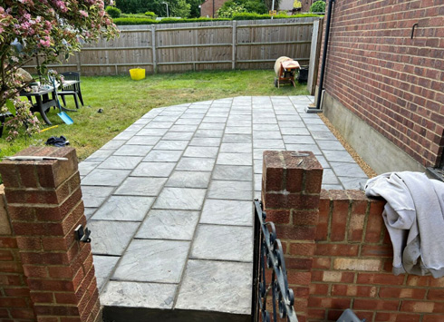 Patio area featuring large gray paving stones, surrounded by a grassy yard and brick walls, with a few outdoor furniture items visible in the background.