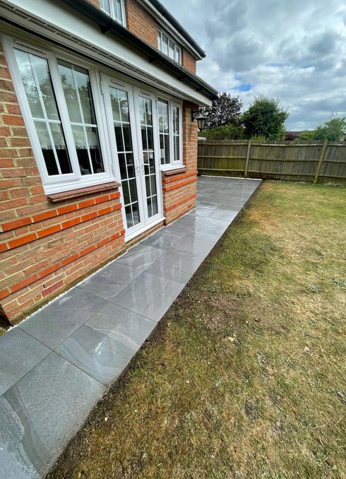 A newly laid gray stone patio extending from a brick house with large windows, surrounded by grass and a wooden fence. Perfect for outdoor living spaces and garden designs.