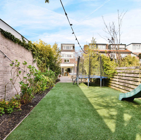 A spacious backyard featuring lush green artificial grass, a trampoline, and a slide, surrounded by shrubs and trees. The scene is bright and inviting, perfect for outdoor family activities.