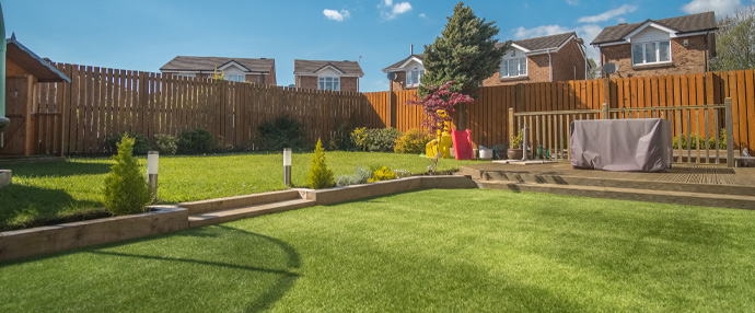 A sunny backyard garden featuring well-maintained grass, a wooden deck, planters, and a wooden fence, with houses in the background.