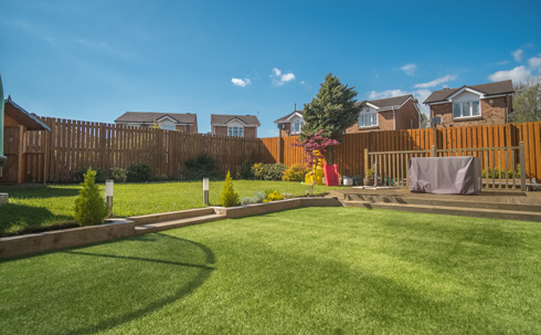 a sunny residential garden with manicured lawn, wooden fence, and houses in the background