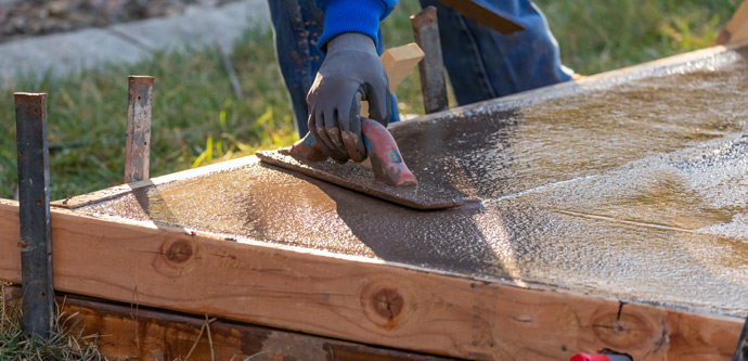 A person wearing gloves smoothing wet concrete with a trowel in a wooden frame on a grassy area.