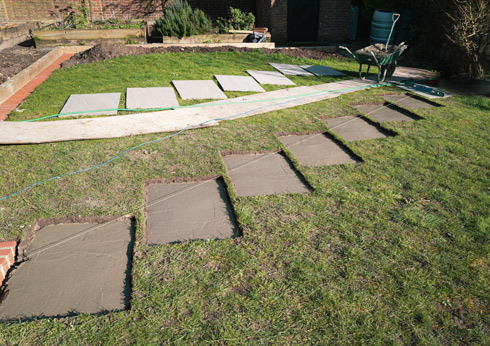 Stepping stones being laid in a garden, with a wheelbarrow and tools in the background on a sunny day.