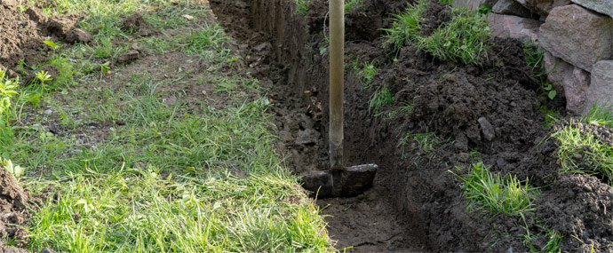 A shovel is positioned in a freshly dug trench surrounded by grass and soil.