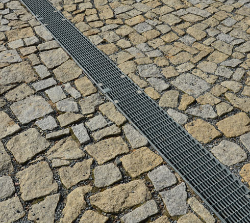 A close-up view of a textured stone pavement featuring irregularly shaped stones and a metallic drainage grate running along the center of the path.