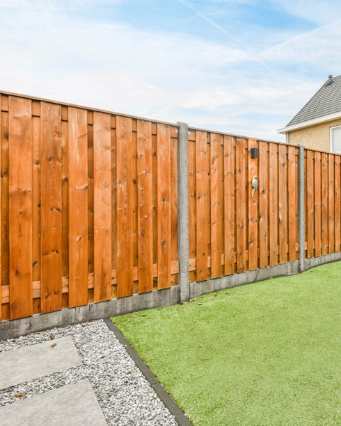 Wooden fence with a concrete base and a small green lawn area, featuring stepping stones and gravel on the side, under a blue sky.