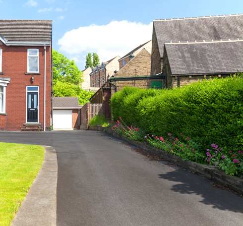 residential driveway leading to a brick house, surrounded by green foliage and flowers, with a clear blue sky in the background