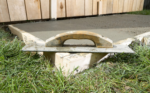 A close-up of a trowel resting on freshly poured concrete in a wooden frame, surrounded by grass, demonstrating concrete finishing techniques.