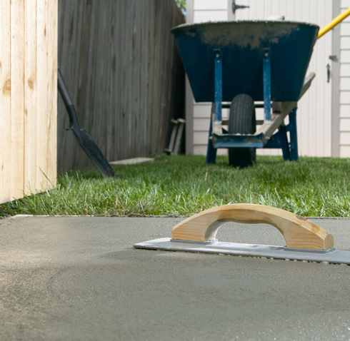 A freshly smoothed concrete surface with a trowel in the foreground, and a wheelbarrow positioned on the grass in the background, indicating a home construction or landscaping project.