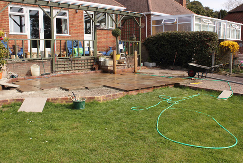 A partially constructed patio area in a residential backyard, featuring laid stone slabs, gardening tools, a wheelbarrow, and a garden hose, surrounded by green grass and landscaping.