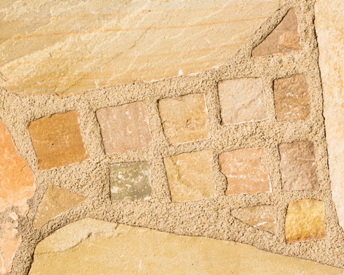 A textured stonework surface featuring various shapes and colors of stones arranged in a pattern, showcasing natural hues of beige, brown, and gray, suitable for outdoor paving.