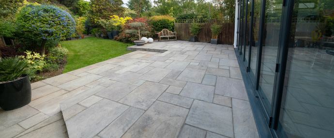 Modern outdoor patio with grey stone paving, surrounded by lush greenery and colorful plants, showcasing a peaceful garden space.