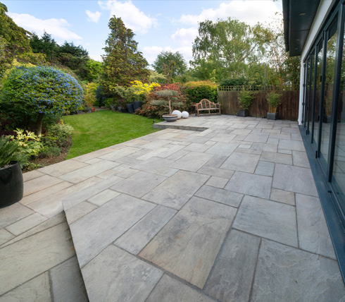 A spacious outdoor patio featuring grey stone tiles, surrounded by a lush garden with various plants, trees, and decorative elements, showcasing a tranquil landscape ideal for relaxation.