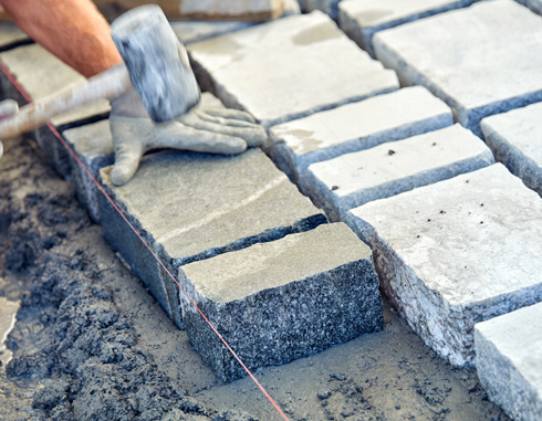 person laying stone pavers on a gravel surface, using a hammer and wearing work gloves to arrange the stones evenly