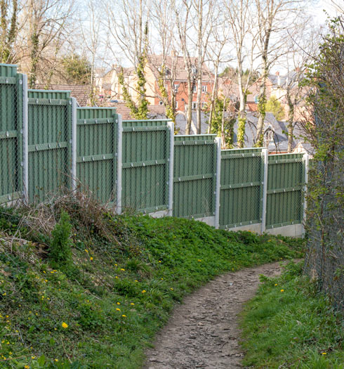 A narrow dirt path bordered by green fencing and trees, leading through a grassy area towards houses in the background.