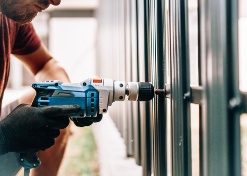 Person using a power drill to fasten a fence panel, wearing gloves and focusing on the task.