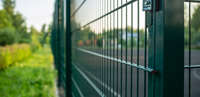 A close-up view of a green metal fence surrounding a sports court, with plants and trees visible in the background, highlighting the outdoor area.