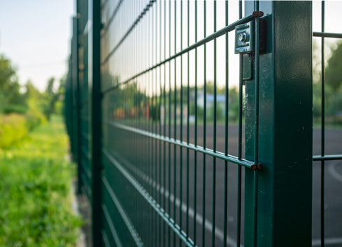 Close-up view of a green metal fence with a latch, set against a blurred background of grass and a sports court.