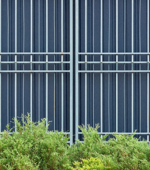 A blue metal fence with vertical bars in a geometric pattern, surrounded by green shrubs and foliage. Ideal for modern architecture.