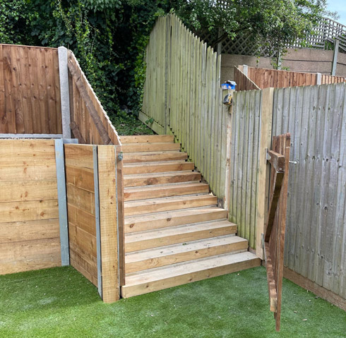 Wooden stairs leading to an outdoor area, flanked by wooden fences, with grassy ground and greenery nearby.